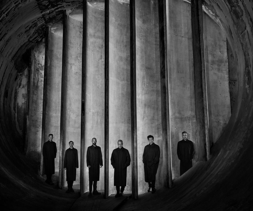 Muzycy zespołu Rammstein w ciemnych płaszczach na tle betonowej konstrukcji. O zespole przeczytasz na EskaRock.