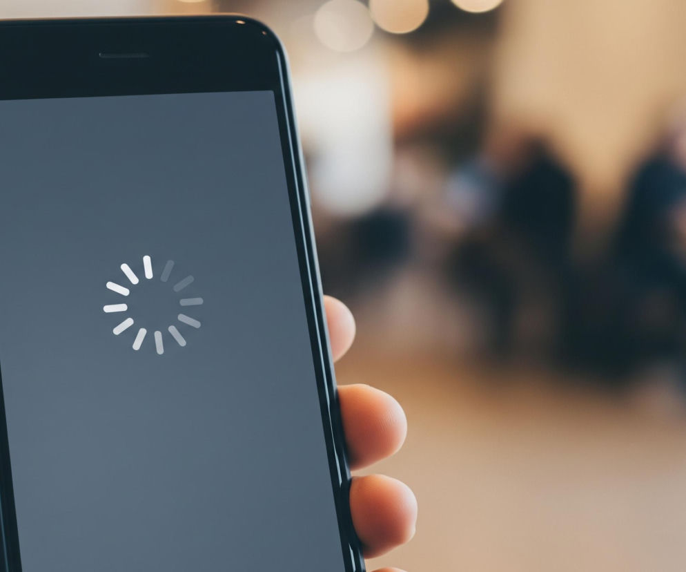 Zbliżenie na dłoń trzymającą czarny smartfon, którego ekran jest wygaszony lub wyświetla szare tło z białym, obracającym się wskaźnikiem ładowania. Palce dłoni są lekko zgięte, obejmując boki urządzenia. W tle widoczne są rozmyte kształty i kolory, sugerujące obecność innych osób i obiektów w niewyraźnym otoczeniu, o ciepłych, beżowych i ciemnych barwach.