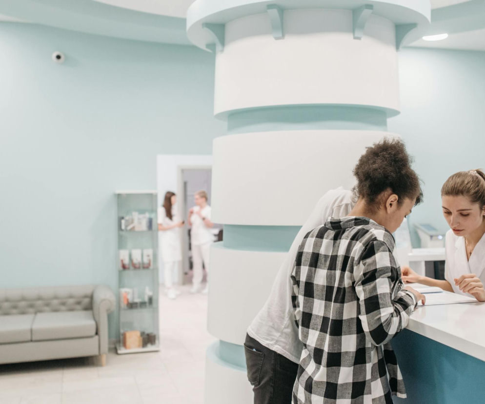 Pacjentka rejestruje się w placówce medycznej, wypełniając dokumenty przy stanowisku obsługi, podczas gdy obok stoi wspierający ją mężczyzna. W tle widoczni są inni pracownicy i wygodna poczekalnia. Na Poradnik Zdrowie znajdziesz informacje o przygotowaniu do biopsji.