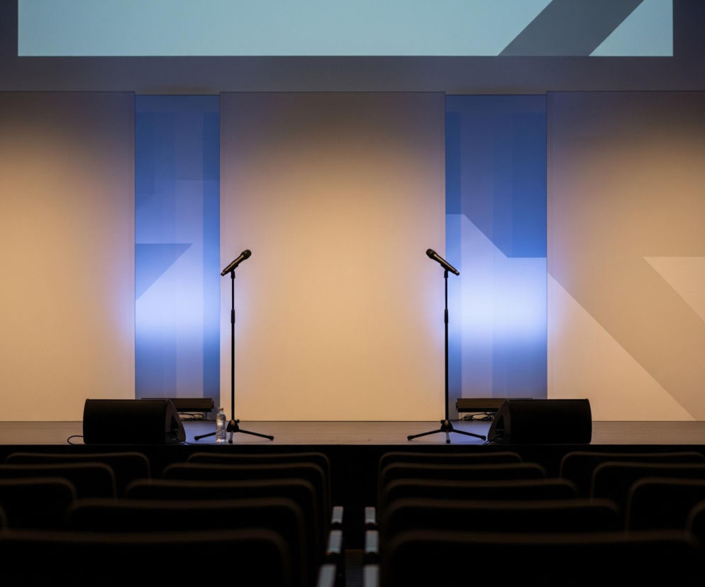 Centralnym punktem obrazu jest scena oświetlona ciepłym, pomarańczowo-żółtym światłem w środku i niebieskim po bokach, na której stoją dwa czarne mikrofony na statywach. Za sceną znajduje się ściana podzielona na prostokątne panele w odcieniach szarości i błękitu, z geometrycznymi wzorami przypominającymi strzałki. Przed sceną, na pierwszym planie, widoczne są ciemne, puste rzędy foteli, sugerujące audytorium lub salę konferencyjną. Całość tworzy nowoczesny i uporządkowany wygląd, z wyraźnym podziałem na część dla występujących i dla publiczności.