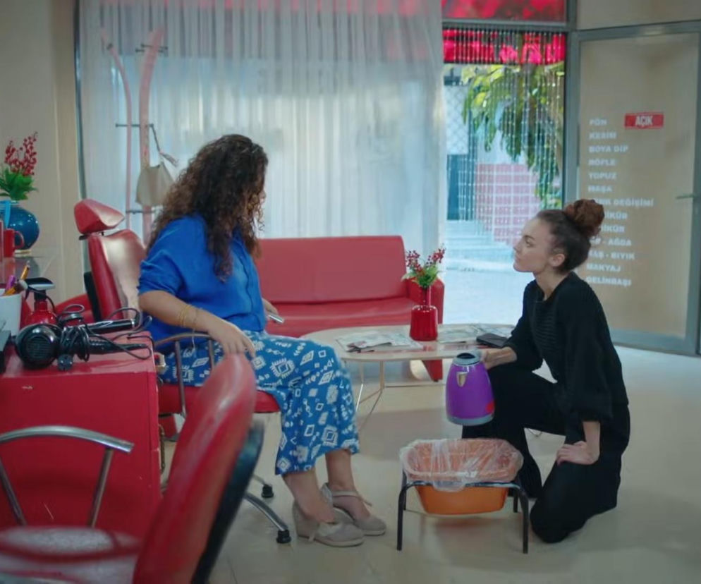 Dwie kobiety w salonie fryzjerskim; jedna siedzi, druga klęczy, trzymając czajnik nad miską do pedicure. Kobieta w niebieskiej bluzce to Nana, a klęcząca to Cansel. Scena z serialu Dziedzictwo ukazująca odwet Nany. Więcej o ich losach na Super Seriale.