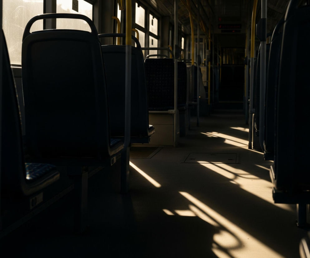 Wnętrze pojazdu transportu publicznego, prawdopodobnie autobusu lub tramwaju, przedstawione jest z perspektywy środkowego przejścia, skierowane ku tyłowi. Pomarańczowe światło słoneczne wpada przez okna po lewej stronie, tworząc na podłodze długie, ukośne pasy światła i wyraźne cienie siedzeń oraz pionowych rurek. Większość siedzeń po obu stronach przejścia jest w cieniu, widoczne są jedynie ich ciemne, tapicerowane oparcia i zagłówki, z nielicznymi prześwitami na tapicerce siedzisk. Wzdłuż sufitu ciągną się żółte poręcze, a w tle widać ciemne, niewyraźne wnętrze pojazdu.