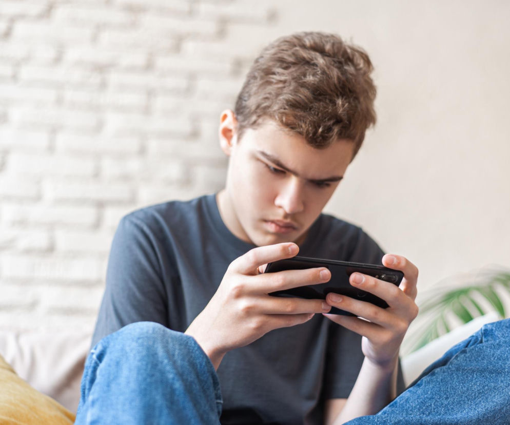 Zamyślony młody chłopak z brązowymi włosami i w jeansach, siedzący na beżowej kanapie, patrzy na ekran smartfona. Obraz symbolizuje problem samodiagnozy ADHD i autyzmu po obejrzeniu filmików z TikToka. O tym, jak łatwo o pomyłkę, przeczytasz na Poradnik Zdrowie.