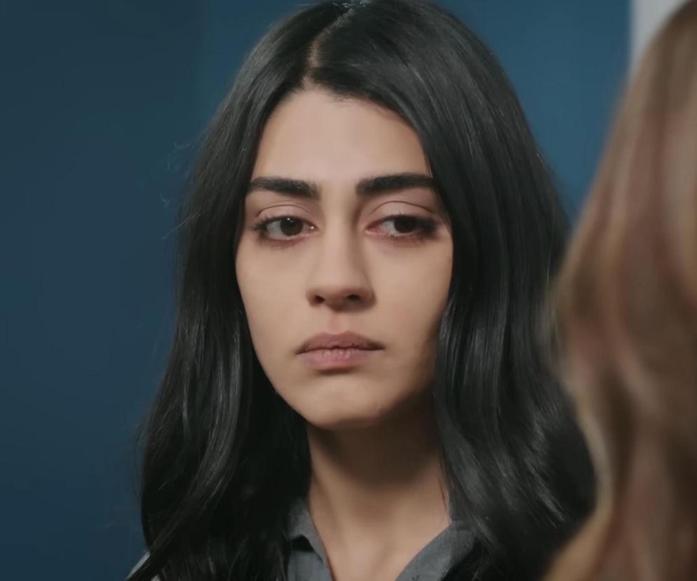 Hancer z tureckiej telenoweli „Panna młoda” z długimi, ciemnymi włosami, spoglądająca przed siebie z lekko zamyśloną miną. Widoczne tło to jednolita, ciemnoniebieska ściana. Zdjęcie przedstawia główną bohaterkę, o której więcej przeczytasz na portalu Super Seriale.