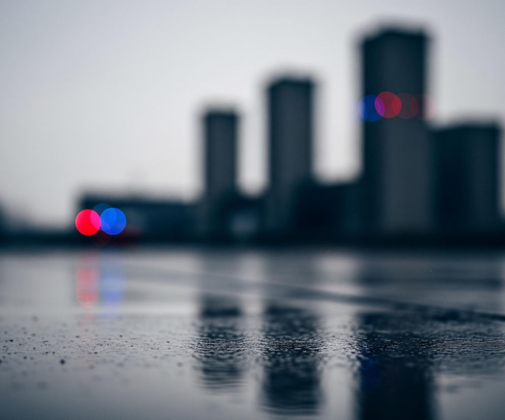 Widoczna jest mokra powierzchnia, prawdopodobnie asfaltowa lub betonowa, z licznymi kroplami wody, tworząca odbicia. Na pierwszym planie, w lewym i prawym dolnym rogu, ta powierzchnia jest ostra, a w głębi obrazu rozmywa się. Na horyzoncie, w tle, znajduje się szereg rozmytych, ciemnych sylwetek budynków, przypominających wieżowce, a po prawej stronie widać niewyraźne, czerwone i niebieskie okrągłe światła, które odbijają się także na mokrej nawierzchni. Cały obraz utrzymany jest w zimnych, szarych i niebieskich tonach, a niebo jest jasne i jednolite, bez wyraźnych chmur.