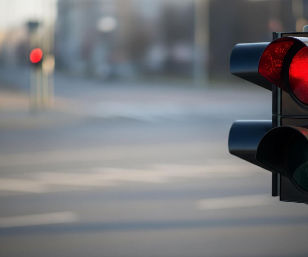 Po prawej stronie zdjęcia znajduje się pionowy sygnalizator świetlny z dwoma otworami. Górne światło świeci na czerwono, rzucając intensywną barwę, podczas gdy dolne jest zgaszone i czarne. Sygnalizator jest przymocowany do jasnoszarego słupa, który rozciąga się w pionie. W tle, po lewej stronie, widać rozmyty obraz ulicy z innym, odległym czerwonym światłem oraz niewyraźnymi elementami miejskiego krajobrazu.