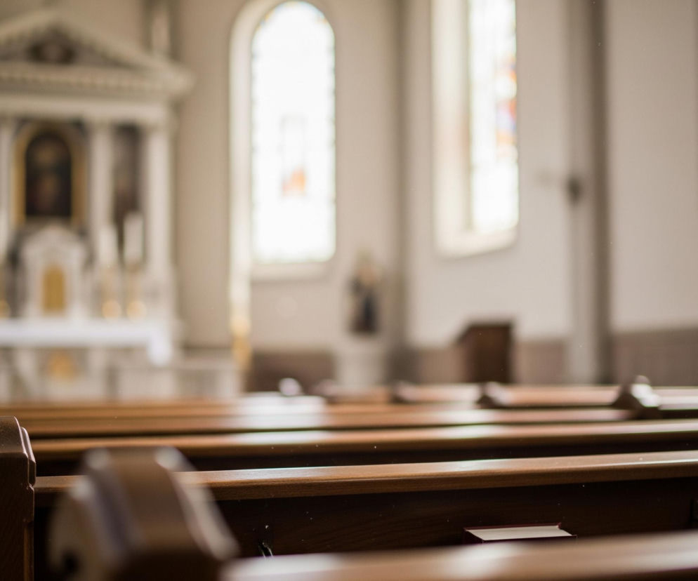 Drewniane ławki kościelne w brązowym kolorze, z rzeźbionymi zakończeniami, biegną ukośnie od prawej do lewej strony w dolnej części kadru. Z przodu w jednej z ławek leży mała, czerwona książeczka. Tło jest rozmyte, ukazując jaśniejsze wnętrze, gdzie w oddali widać ołtarz z kolumnami oraz dwa wysokie, łukowate okna, z których jedno zawiera witraż w barwach niebieskiego i pomarańczowego.