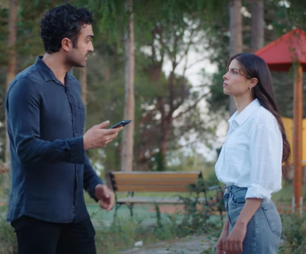 Kadr z serialu Dziedzictwo, odcinek 897, przedstawia Ferita z telefonem i prokuratorkę Leylę w parku, gdzie Ayse odkrywa ich spotkanie. Ferit ma ciemne kręcone włosy i brodę, ubrany jest w granatową koszulę i czarne spodnie. Leyla ma długie, ciemne włosy, ubrana jest w białą koszulę i jasne jeansy. W tle widać ławkę i elementy placu zabaw. Więcej o ich losach na Super Seriale.