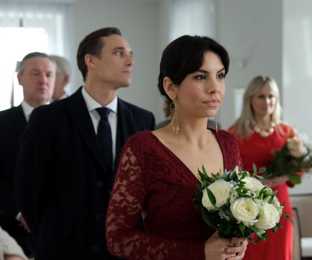 Aneta (Ilona Janyst) w bordowej koronkowej sukience, z bukietem białych róż, obserwuje ceremonię ślubną Kasi i Mariusza w M jak miłość. Obok niej stoi pan młody Mariusz (Mateusz Mosiewicz) w garniturze. O tym, jak Aneta uratuje ten ślub, przeczytasz na stronie Super Seriale.