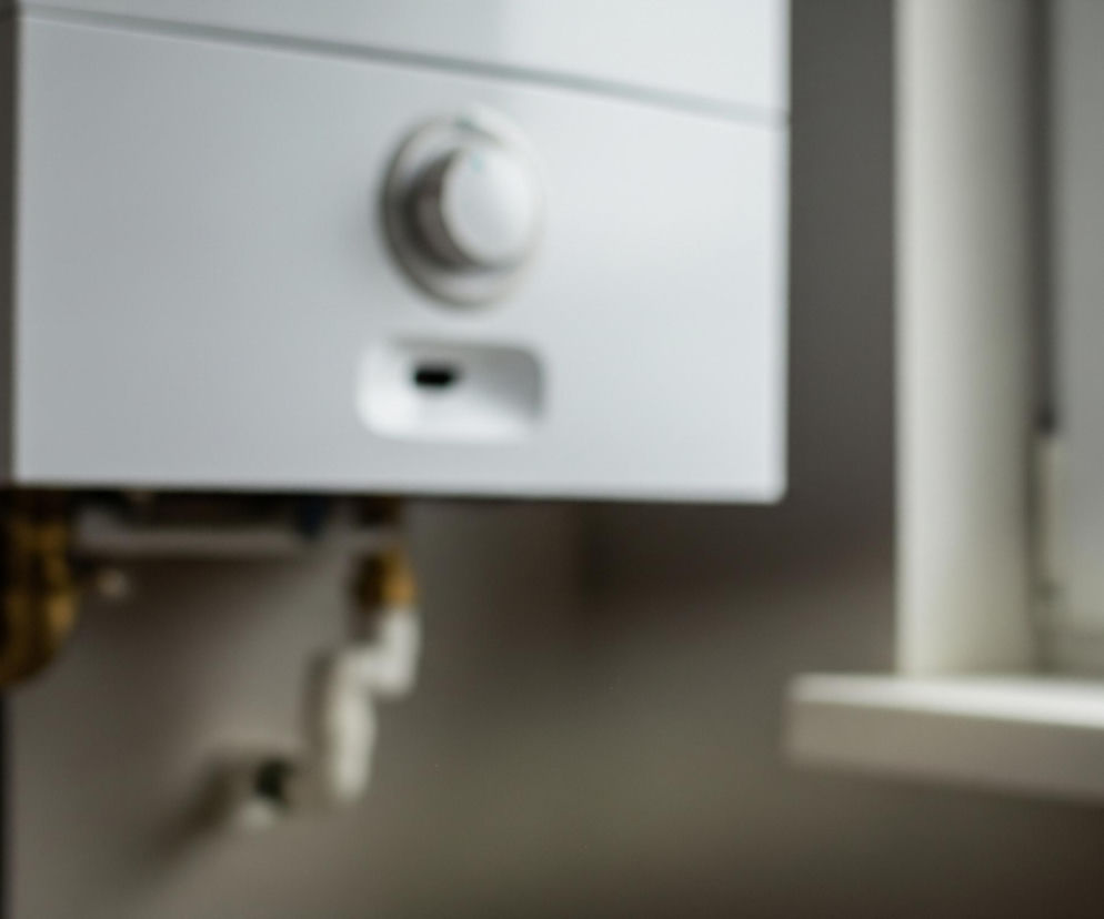 Biały, prostokątny kocioł gazowy z okrągłym pokrętłem i podłużnym wlotem w centralnej części, dominuje po lewej stronie kadru. Poniżej kotła widoczne są mosiężne i białe rury instalacyjne, biegnące poziomo i pionowo na ciemnej ścianie. Po prawej stronie, w tle, znajduje się fragment białego okna z parapetem, rozmytego i jaśniejszego.