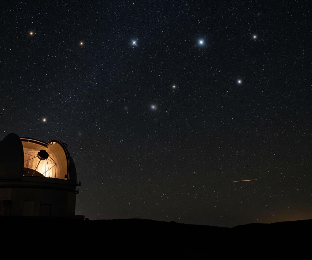 Duża kopuła obserwatorium astronomicznego z otwartą szczeliną w górnej części, ukazującą jasno oświetlone wnętrze z fragmentem teleskopu, dominuje w lewej dolnej części kadru. Konstrukcja teleskopu jest widoczna jako ciemne pręty i okrągłe elementy. Obserwatorium stoi na ciemnym, sylwetkowym wzniesieniu terenu, które rozciąga się przez cały dół obrazu. Nad kopułą i horyzontem rozciąga się ciemne, usiane mnóstwem małych, białych i żółtawych punktów niebo, tworząc widok rozgwieżdżonej nocy. Kilka jaśniejszych gwiazd tworzy wyraźne konstelacje, a jedna z nich przypomina Małą Niedźwiedzicę. Na prawo od kopuły, na niebie, widoczna jest jasna, pomarańczowa smuga światła.