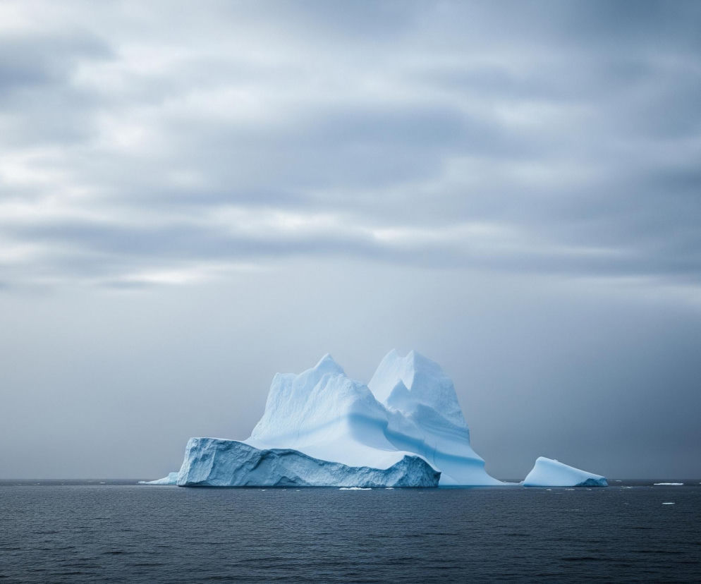 W centrum obrazu znajduje się duża góra lodowa o nieregularnym kształcie, składająca się z kilku poszarpanych szczytów i stromych ścian, lśniąca w odcieniach bieli i błękitu. Jej podstawa zanurza się w ciemnoszarych, lekko pofalowanych wodach morza, które rozciąga się od dolnej krawędzi obrazu po horyzont. Ponad górami lodowymi i morzem widoczne jest zachmurzone niebo, o zmiennej barwie od jasnoszarej po lewej stronie, przechodzącej w ciemniejszy, grafitowy odcień po prawej. Drobne kawałki lodu unoszą się na wodzie w oddali, zarówno po lewej, jak i po prawej stronie głównej bryły.