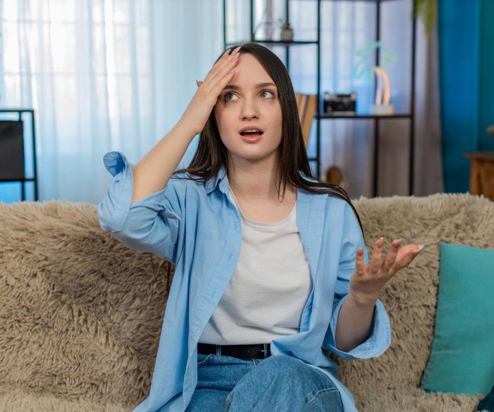 Młoda kobieta w jasnoniebieskiej koszuli i białym T-shircie, siedząca na beżowej, puszystej sofie. Gestem dłoni przy czole wyraża frustrację lub zapomnienie, a jej otwarte usta i spojrzenie w górę sugerują próbę przypomnienia sobie czegoś. Scena ilustruje codzienne problemy z pamięcią, o których więcej przeczytasz na Poradnik Zdrowie.