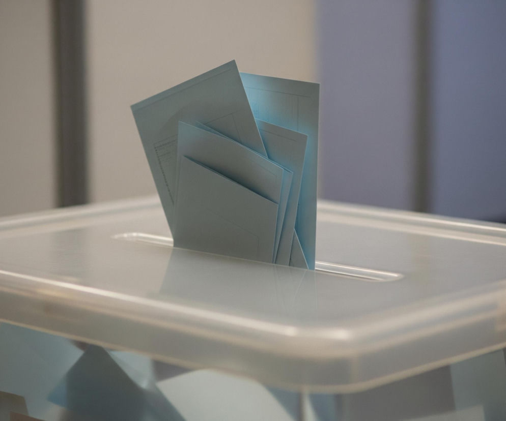 W centrum zdjęcia znajduje się przezroczysta urna wyborcza wykonana z jasnego, półprzezroczystego plastiku. Na jej górnej powierzchni, pośrodku, widoczny jest podłużny otwór, przez który wystaje kilka złożonych kartek papieru w kolorze jasnoniebieskim lub błękitnym. Kartki są częściowo włożone do urny, z ich górnymi krawędziami skierowanymi ku górze i nieco rozchylonymi. Wnętrze urny również zawiera zagięte kartki o podobnym kolorze, tworzące nieregularną masę. Tło jest rozmyte i utrzymane w odcieniach szarości, z jaśniejszymi elementami po lewej stronie i ciemniejszymi po prawej.