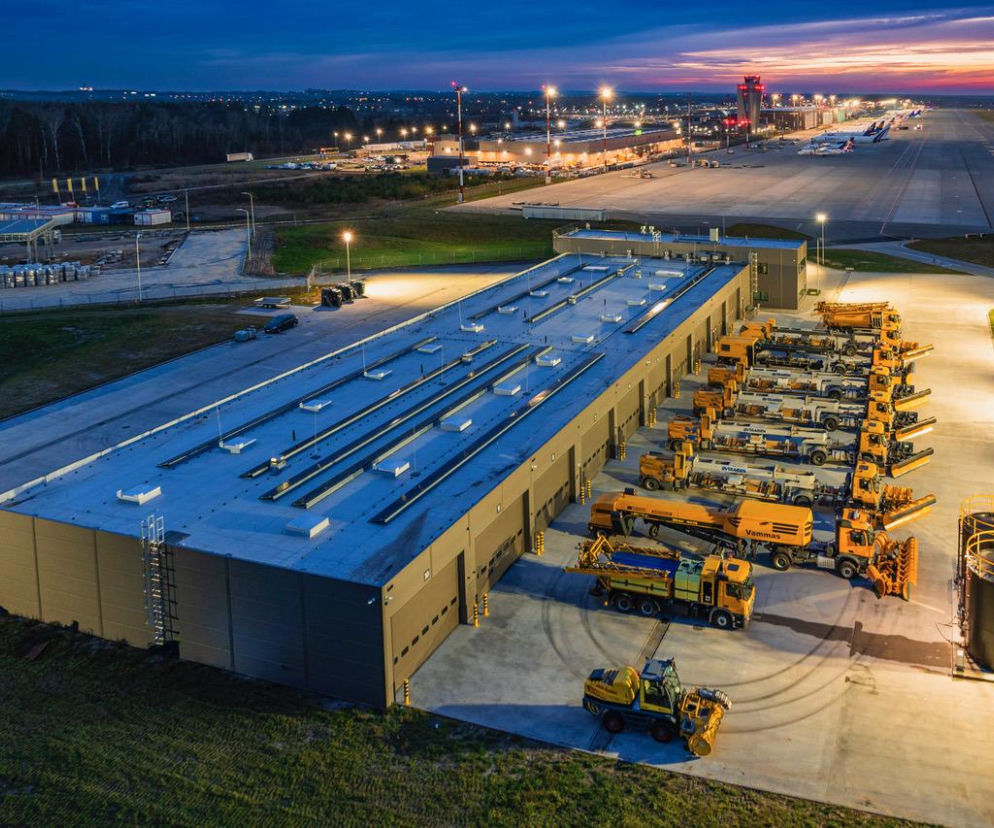 Nowoczesna baza Działu Eksploatacji i Transportu Katowice Airport z halą garażową, gdzie stacjonują żółte oczyszczarki lotniskowe. Widoczne pasy startowe i kontrolna wieża lotniska o zmierzchu. Więcej o inwestycjach lotniskowych przeczytasz na Architektura Murator Plus.