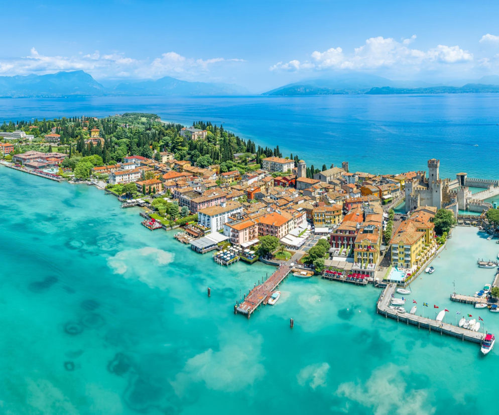 Malownicze Sirmione nad jeziorem Garda z zamkiem Scaligerich i kolorowymi kamienicami otoczonymi turkusową wodą, podkreślające urok miejsca opisywanego na naszym portalu. W tle góry i bezkresne błękitne niebo.