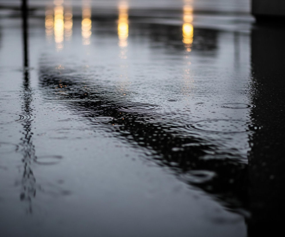 Powierzchnia pokryta wodą, prawdopodobnie betonowa lub asfaltowa, odbija światło. Widoczne są liczne drobne krople deszczu i koncentryczne kręgi fal tworzone przez spadające krople, zwłaszcza w dolnej części kadru. W górnej części obrazu, na horyzoncie, rozmyte, pionowe smugi jaskrawożółtego światła odbijają się od mokrej powierzchni, kontrastując z ciemnymi, rozmyślonymi odbiciami w tle, które przypominają słupy lub konstrukcje. Prawa strona obrazu jest znacznie ciemniejsza, z mniej widocznymi detalami, co tworzy głęboki cień lub obszar bez oświetlenia.