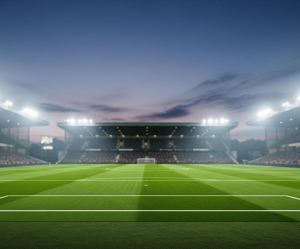 Jasnozielone boisko piłkarskie z białymi liniami wyznacza strefy gry, biegnąc przez dolną część kadru. Trawa ułożona jest w pasy o naprzemiennych odcieniach zieleni, jaśniejsze bliżej widza, a ciemniejsze w oddali. Po obu stronach boiska widać trybuny wypełnione rozmazanymi postaciami kibiców. Nad trybunami wznoszą się maszty oświetleniowe z rzędami jasnych reflektorów, oświetlających całe boisko. Niebo jest ciemnoniebieskie, z delikatnymi, szarymi chmurami, sugerującymi wieczór lub zmierzch.