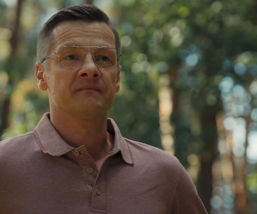 Zbliżenie na twarz Piotrka Zduńskiego (Marcin Mroczek) w okularach, ubranego w różową koszulkę polo, na tle niewyraźnych drzew. Zdjęcie ilustruje emocje związane ze zniknięciem Pawła, o czym przeczytasz więcej na portalu Super Seriale.