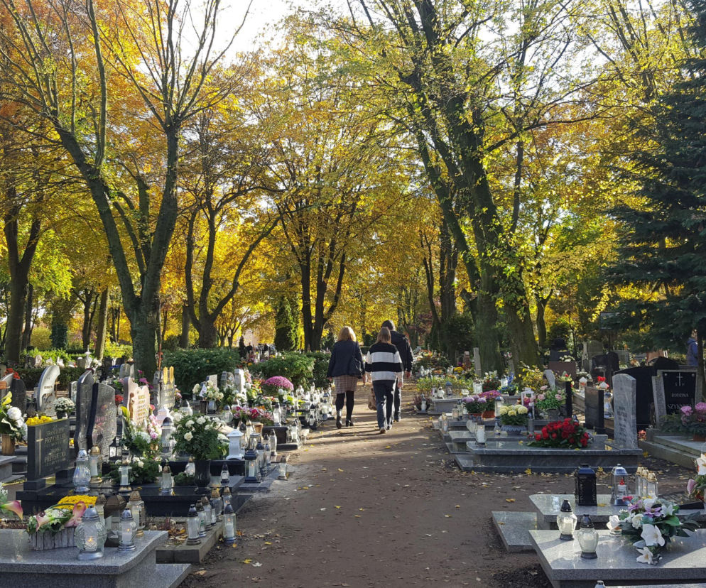 Ludzie idący alejką cmentarza wśród grobów z kwiatami i zniczami. O zmianach w Lesznie przeczytasz na Eska Leszno.