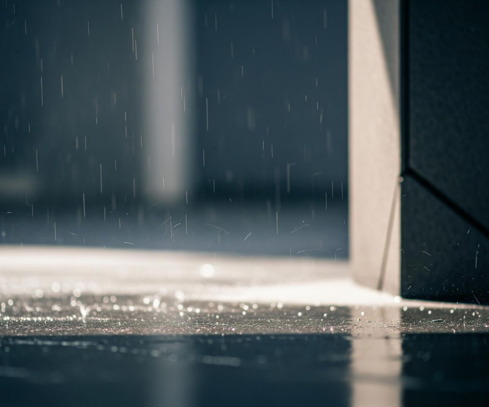 Deszcz pada na ciemną, błyszczącą powierzchnię, która odbija światło w postaci jasnych punktów i smug. Po prawej stronie widać fragment jasnej, beżowej ściany o złożonej, kanciastej strukturze, z ostrym przejściem do ciemniejszego, nieokreślonego tła. Całość utrzymana jest w chłodnej palecie barw, od błękitno-szarych do niemal czarnych tonów, z wyraźnym, kontrastującym oświetleniem kropel deszczu i części powierzchni.