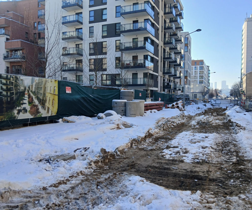 Zimowa sceneria placu budowy na ul. Okrzei w Warszawie, gdzie leży śnieg, widoczne są materiały budowlane i tablica informacyjna z wizualizacją przyszłej ulicy. Więcej o przebudowie tej części stolicy można przeczytać na Architektura Murator Plus.