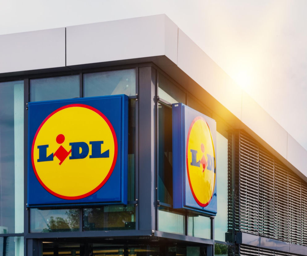 Elewacja sklepu Lidl z widocznym niebiesko-żółtym logo, na tle jasnego nieba. Słońce świeci zza budynku, tworząc jasną poświatę. Temat promocji i wyprzedaży w Lidlu znajdziesz na Super Biznes.