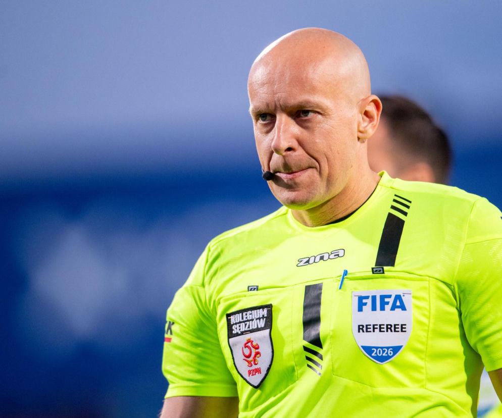 Szymon Marciniak, polski sędzia piłkarski, w zbliżeniu na tle niebieskiego boiska, ubrany w jaskrawozielony strój sędziowski z logo FIFA Referee 2026 i Kolegium Sędziów PZPN. Jego zamyślony wyraz twarzy odzwierciedla kontrowersje wokół decyzji sędziowskich na MŚ 2026, o czym możesz przeczytać na naszym portalu.
