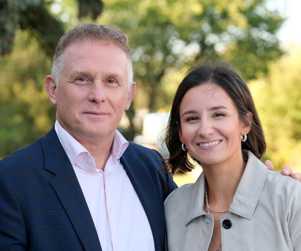 Uśmiechnięta Amanda Mincewicz jako Agnes w beżowej kurtce i Robert Moskwa jako Artur w granatowej marynarce pozują na tle zieleni. Oboje patrzą prosto w obiektyw, symbolizując nadchodzące zmiany w rodzinie Rogowskich w serialu M jak miłość. Więcej o tym na Super Seriale.