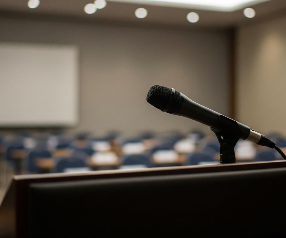 Czarny mikrofon stoi na ciemnobrązowym, skórzanym pulpicie w centrum kadru, skierowany w lewą stronę. Za nim rozciąga się rzędami pusta sala wykładowa z niebieskimi krzesłami i jasnobrązowymi stolikami. W tle widoczny jest duży, biały ekran projekcyjny oraz beżowe ściany i sufit z białymi, okrągłymi światłami. Całość jest słabo oświetlona, z ostrym pierwszym planem i rozmytym tłem.