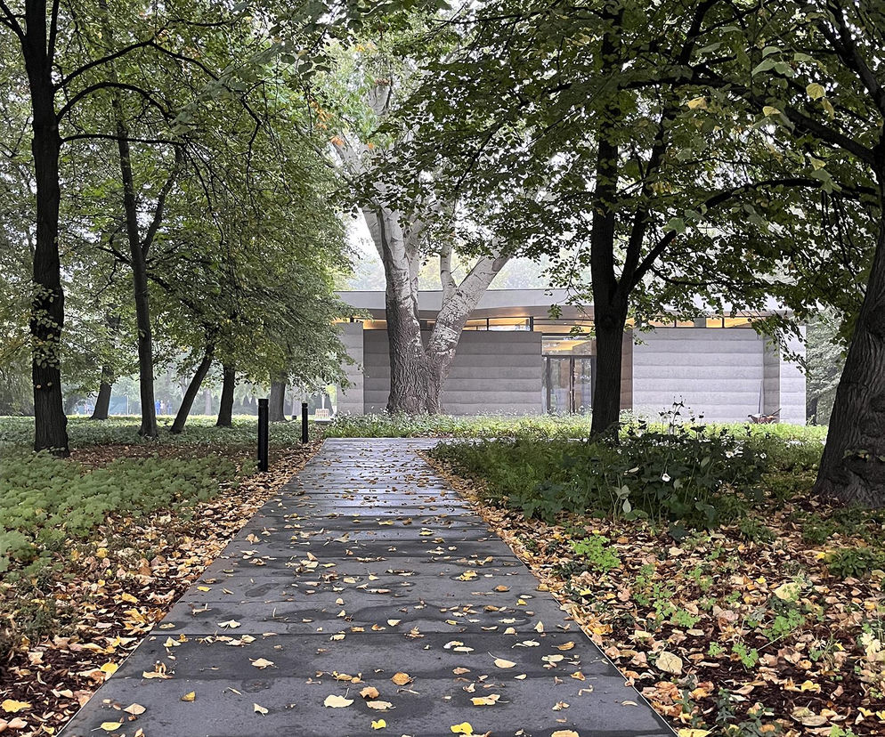 Aleja prowadząca do Izby Pamięci przy Cmentarzu Powstańców Warszawy, projektu Bujnowski Architekci. Ścieżka otoczona jest drzewami i jesiennymi liśćmi, symbolizującymi pamięć o przeszłości. Architektura Murator Plus promuje takie ważne projekty.