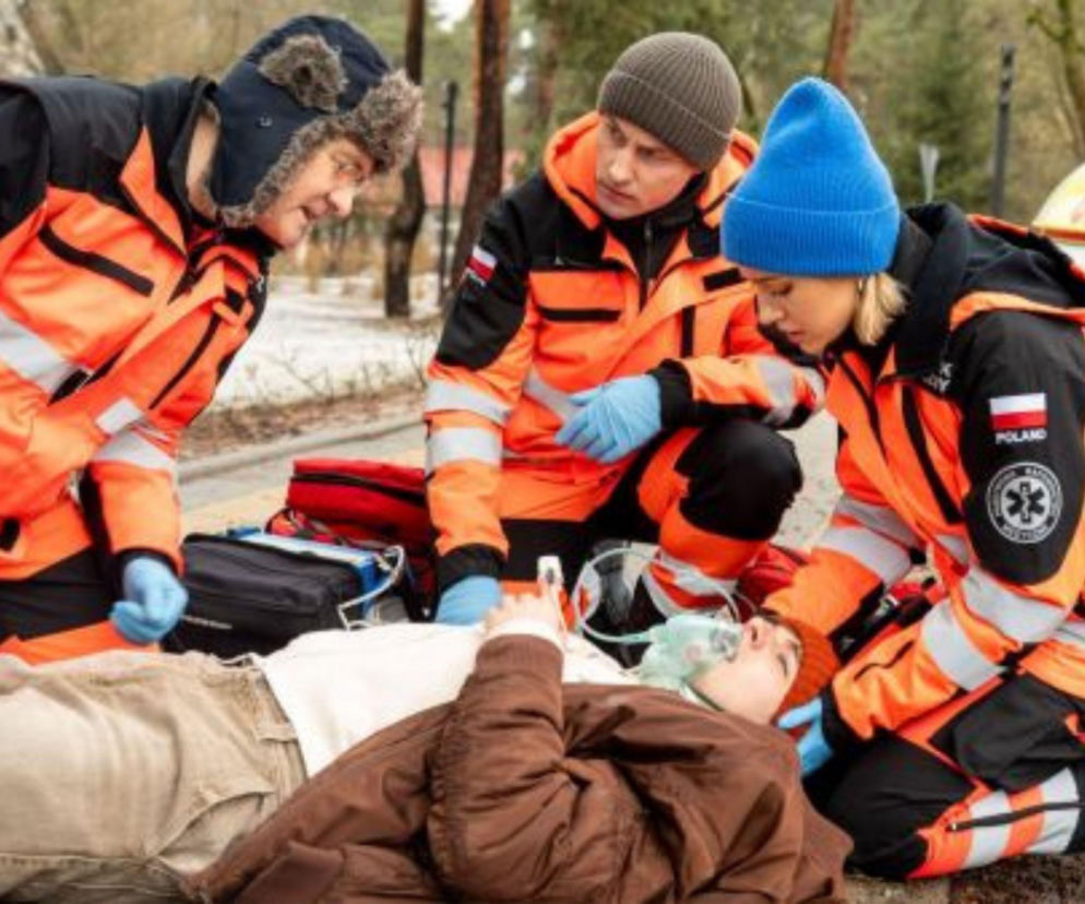 Trzech ratowników medycznych w pomarańczowych uniformach udziela pomocy leżącemu pacjentowi z maską tlenową na twarzy. Ratownicy to Piotr, Alek oraz Martyna. Scena z serialu Na sygnale ukazuje dramatyczne wydarzenia z odcinka 883, o których możesz przeczytać na portalu Super Seriale.