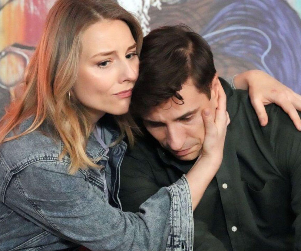 Blondwłosa Grażyna w jeansowej kurtce obejmuje smutnego Tomasza, pocieszając go. Mężczyzna ma ciemne włosy i koszulę, opiera głowę na dłoni kobiety. Na drugim planie widać kolorowe graffiti. Na portalu Super Seriale dowiesz się, czy Kępski wróci do Grażyny.