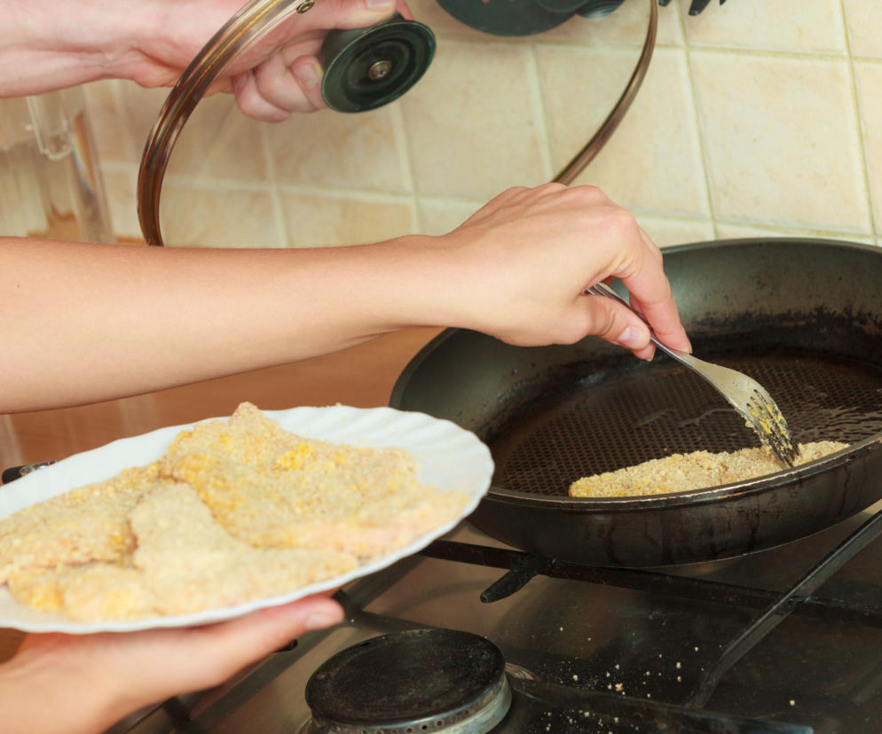 Dłonie smażące panierowane kawałki mięsa na rozgrzanym oleju na patelni, obok talerz z surowym, panierowanym mięsem. Obraz ilustruje niebezpieczeństwa związane z wielokrotnym użyciem oleju, o których możesz przeczytać na Poradnik Zdrowie.