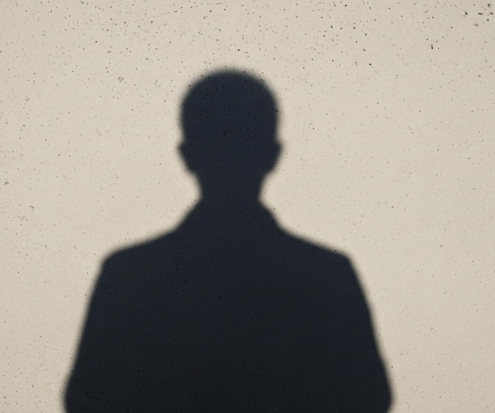 Cień osoby w ciemnym kolorze rzuca się na jasnoszarą, teksturowaną ścianę, zajmując centralną i dolną część kadru. Sylwetka postaci z widoczną głową i ramionami skierowana jest przodem do widza. Po lewej stronie ściany, w pionie, biegnie wąska, ciemniejsza rysa lub szew, rozciągająca się od góry do dołu. Cała powierzchnia ściany jest pokryta małymi, nieregularnymi czarnymi kropkami.