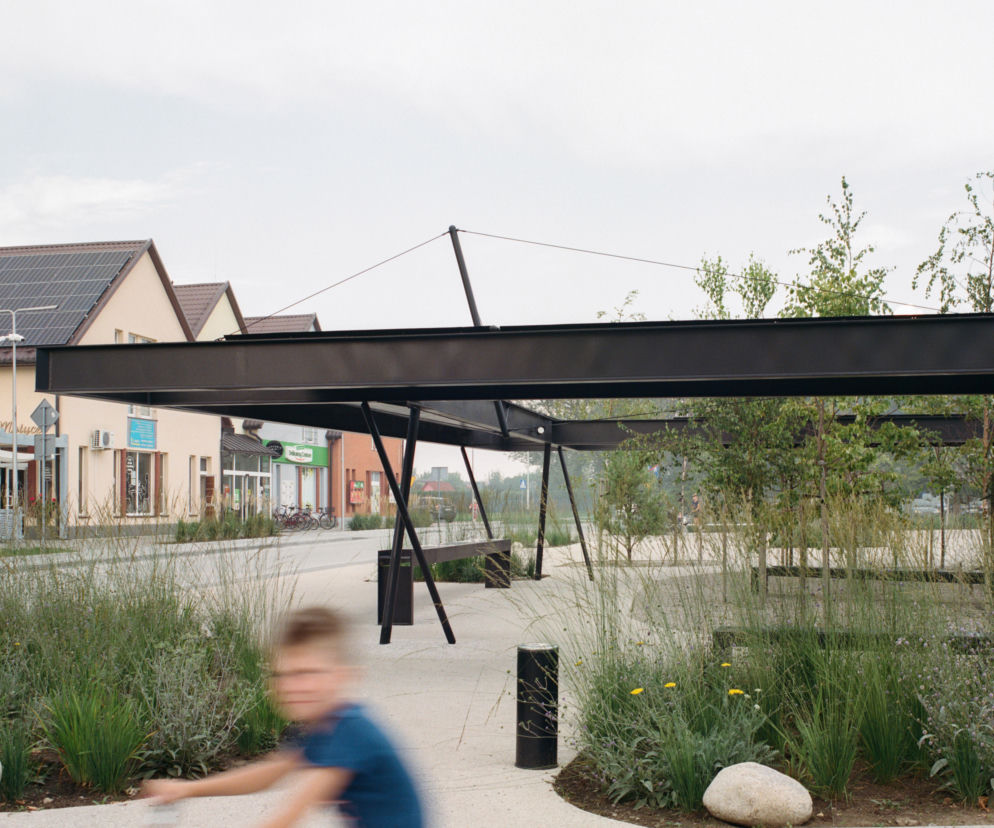 Plac Handlowy w Gorzycach, wyróżniony przez Architektura Murator Plus, prezentuje nowoczesną stalową konstrukcję pawilonu na tle budynków mieszkalnych z panelami fotowoltaicznymi. Przestrzeń jest uporządkowana, z alejkami, zielonymi rabatami, młodymi drzewami i miejscami do siedzenia. Widoczny w ruchu chłopiec i rower podkreślają rekreacyjny charakter miejsca, zaprojektowanego przez Bartosza Bukowskiego i Michała Woźniaka.