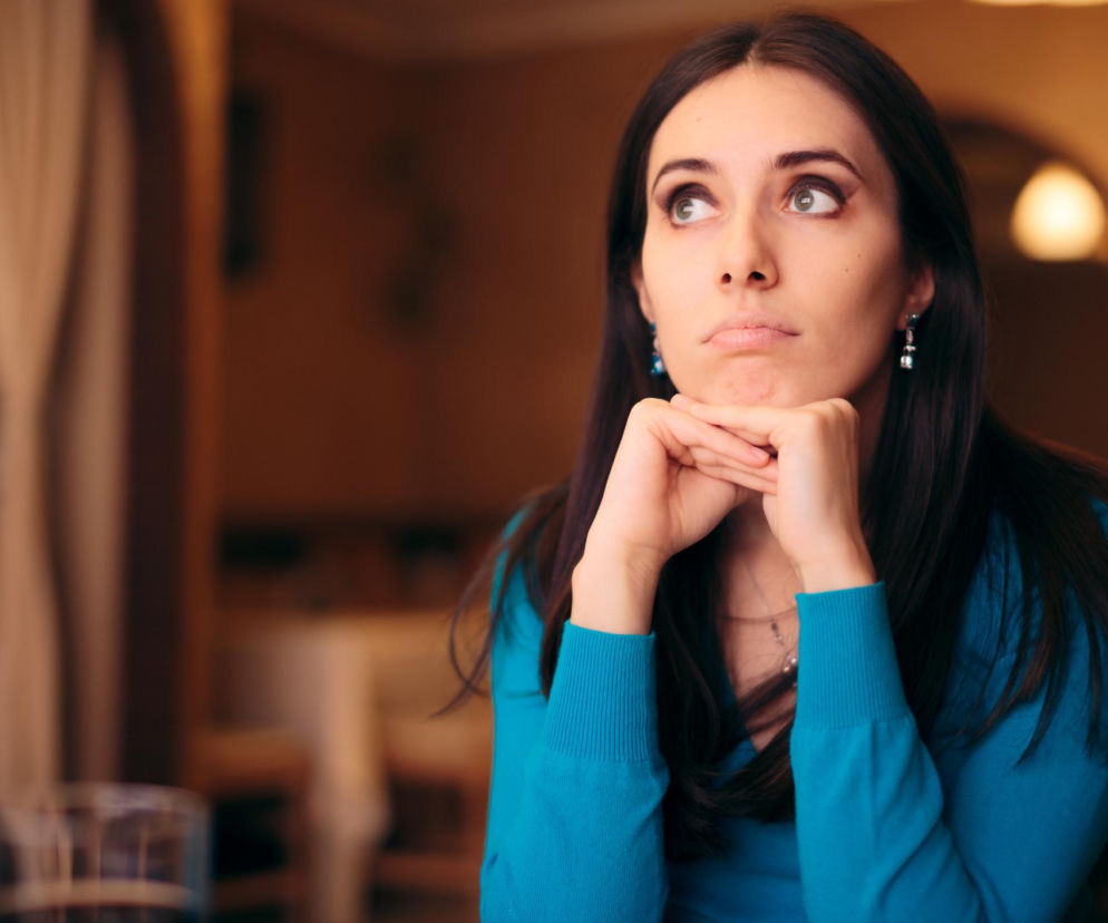 Kobieta z długimi, ciemnymi włosami w turkusowym swetrze, z podpartą brodą, spogląda w górę z zamyśleniem lub znużeniem. Obraz symbolizuje westchnienie jako reakcję na stres lub znużenie, o którym przeczytasz więcej na Poradnik Zdrowie.