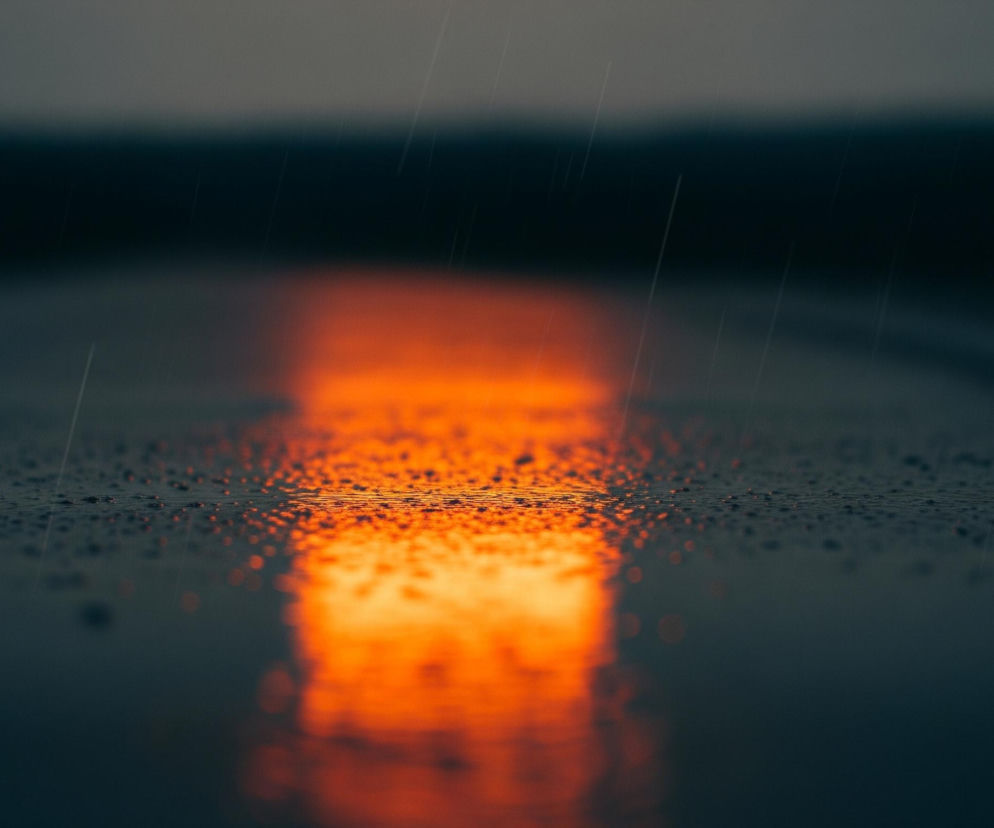Na pierwszym planie widoczna jest pozioma powierzchnia pokryta drobnymi kroplami wody, z wyraźnym odbiciem pomarańczowego światła biegnącego wzdłuż środka kadru. To odbicie rozciąga się od dołu obrazu, rozmywając się ku górze i zmniejszając intensywność barwy. Tło jest rozmyte, z ciemniejszym, niewyraźnym pasmem na środku i jaśniejszym, szarym pasmem u góry. Na całej powierzchni obrazu, od góry do dołu, widoczne są pojedyncze, cienkie linie symbolizujące padający deszcz. Dominują ciemne odcienie szarości i granatu, przełamane intensywną pomarańczą.