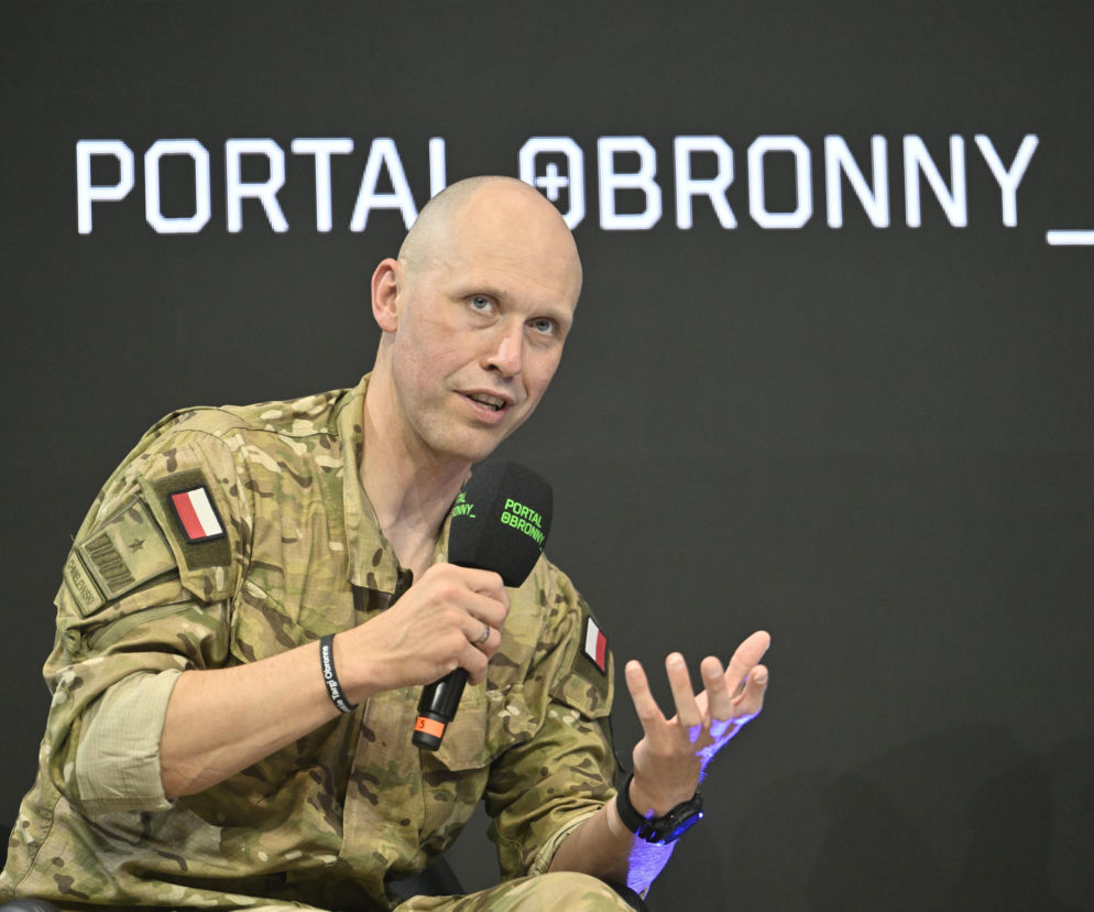 Gen. Mariusz Chmielewski w mundurze moro, podczas prelekcji na Warszawskich Targach Obronnych, trzymający mikrofon z napisem Portal Obronny, z otwartą dłonią w geście mówcy. Za nim widoczny jest logotyp Portal Obronny. O jego pasji do cyberbezpieczeństwa i ekstremalnego biegania przeczytasz więcej na naszym portalu.