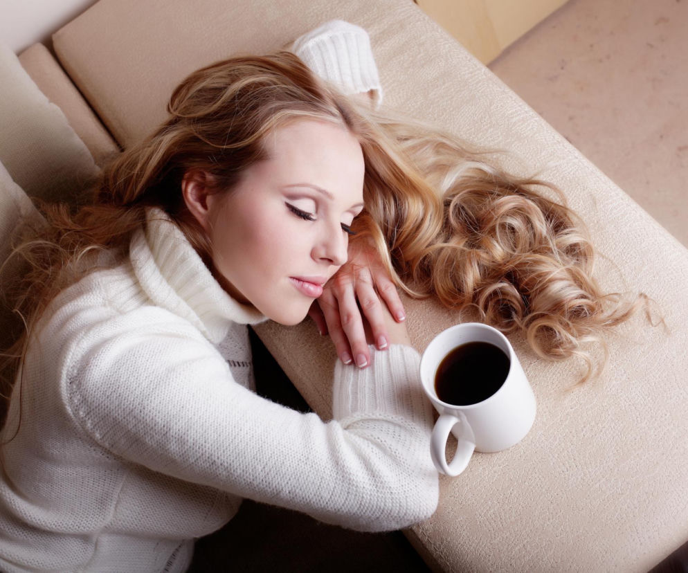 Kobieta w białym swetrze śpi na beżowej sofie obok kubka kawy, demonstrując kawową drzemkę – metodę walki z popołudniowym kryzysem. Dowiedz się więcej o tej technice na Poradnik Zdrowie.
