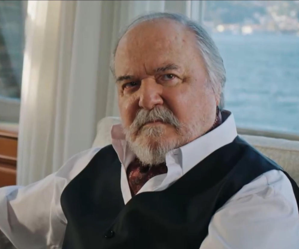 Mężczyzna w średnim wieku, o siwych włosach i brodzie, ubrany w białą koszulę i ciemną kamizelkę. Ma poważny wyraz twarzy. To Çetin Tekindor, aktor wcielający się w postać Halisa Korhana w serialu Złoty chłopak, którego streszczenie odcinków znajdziesz na Super Seriale. W tle widać fragment błękitnego morza.