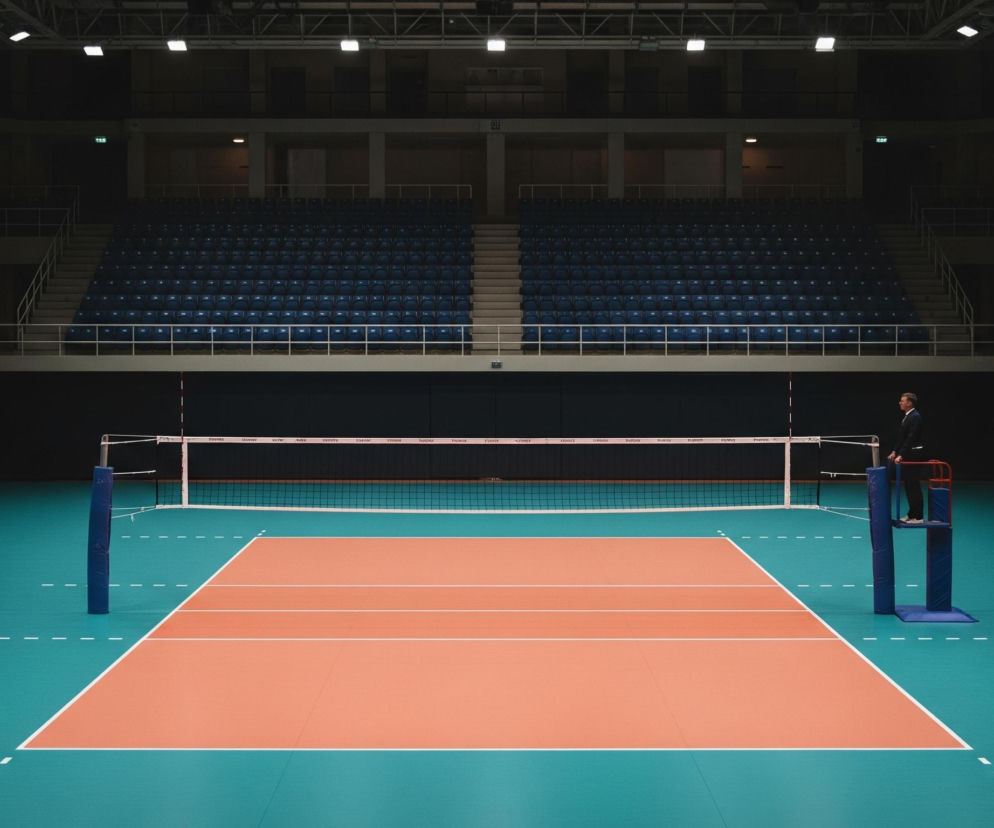 Hala sportowa z pustymi trybunami i boiskiem do siatkówki dominuje na obrazie. W centrum znajduje się siatka do siatkówki rozpięta na słupkach, z centralnym pomarańczowym polem boiska otoczonym turkusową nawierzchnią z białymi liniami. Po prawej stronie siatki stoi mężczyzna w ciemnym garniturze na podwyższeniu sędziowskim. Dookoła boiska widać rzędy granatowych, pustych siedzeń na trybunach, a nad nimi wiszą białe prostokątne lampy oświetlające wnętrze hali.