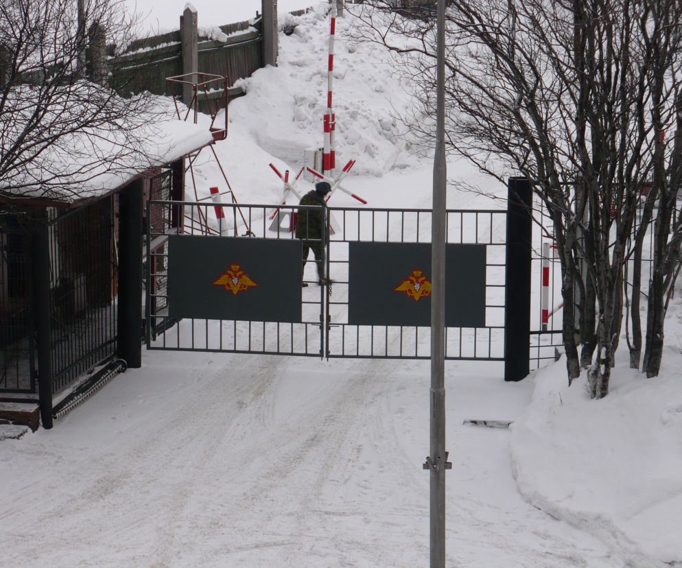 Brama wjazdowa do zamkniętego rosyjskiego miasta Siewieromorsk, otoczona śniegiem i drzewami, z wojskowym emblematem i przechodzącą postacią. ZATO to obszary tajne, o których przeczytasz więcej na Architektura Murator Plus.