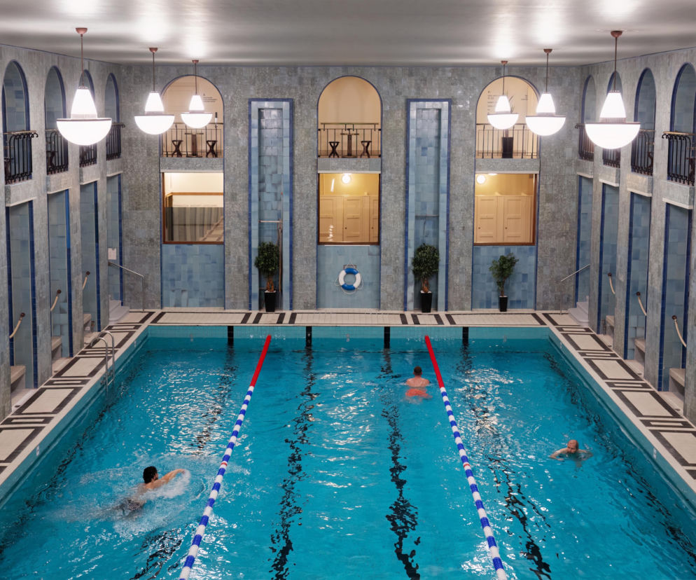 Wnętrze odnowionego basenu Yrjönkatu w Helsinkach, ukazujące jasnoniebieskie płytki i detale architektoniczne w stylu art deco. W basenie pływają trzy osoby, a naokoło widać eleganckie kabiny i balkony z kutymi balustradami, podświetlone kulistymi lampami. O historii renowacji obiektu przeczytasz na Architektura Murator Plus.