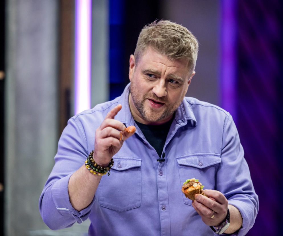Tomasz Jakubiak, kucharz i osobowość telewizyjna, w fioletowej koszuli, trzymający w rękach przygotowane potrawy, jedną wskazując palcem. Zdjęcie oddaje jego naturalność i pasję do gotowania, o których więcej przeczytasz na naszym portalu.