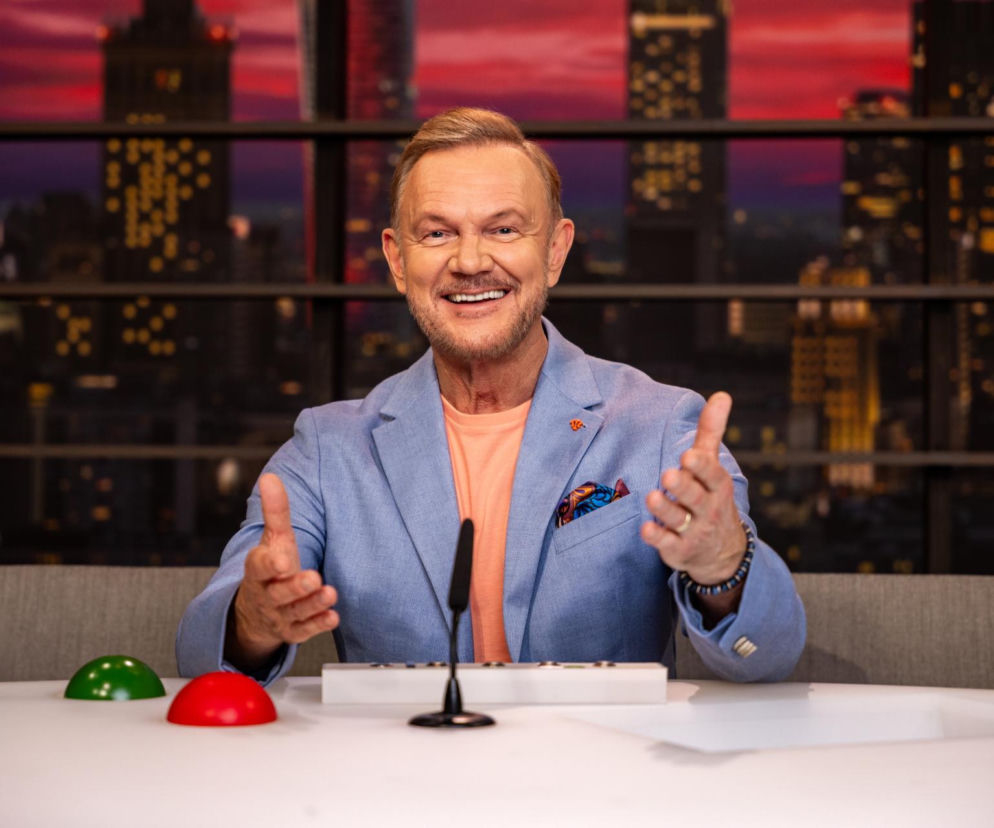 Cezary Pazura uśmiecha się za biurkiem na planie programu LOL. O szczegółach produkcji przeczytasz na SE.