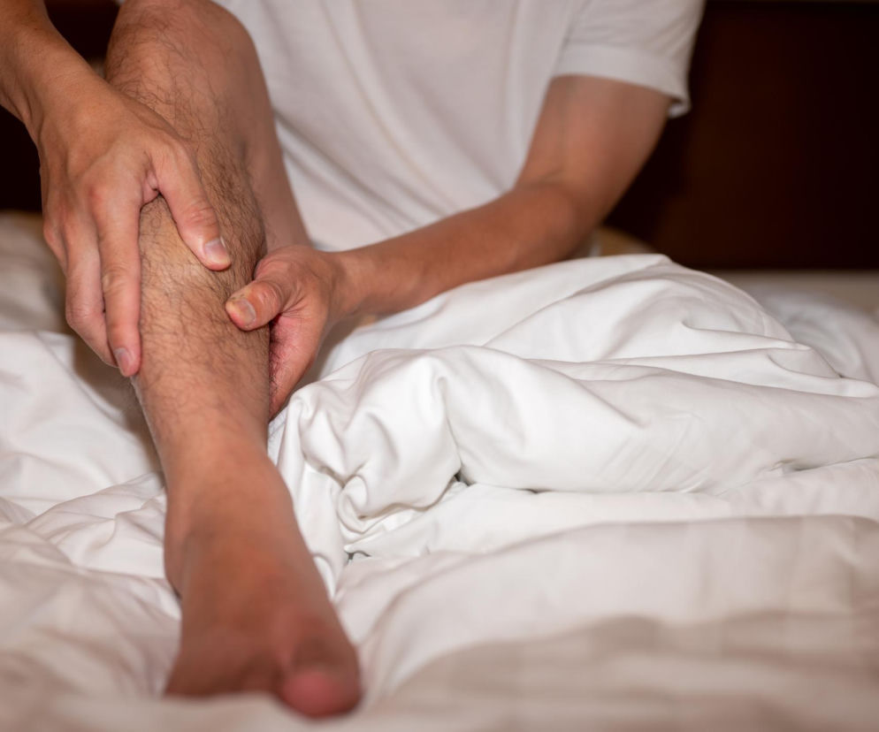 Mężczyzna w białym t-shircie siedzący na łóżku, masujący łydkę, symbolizujący problem niespokojnych nóg i mrowienia. Artykuły na temat przyczyn i leczenia tego schorzenia znajdziesz na Poradnik Zdrowie.