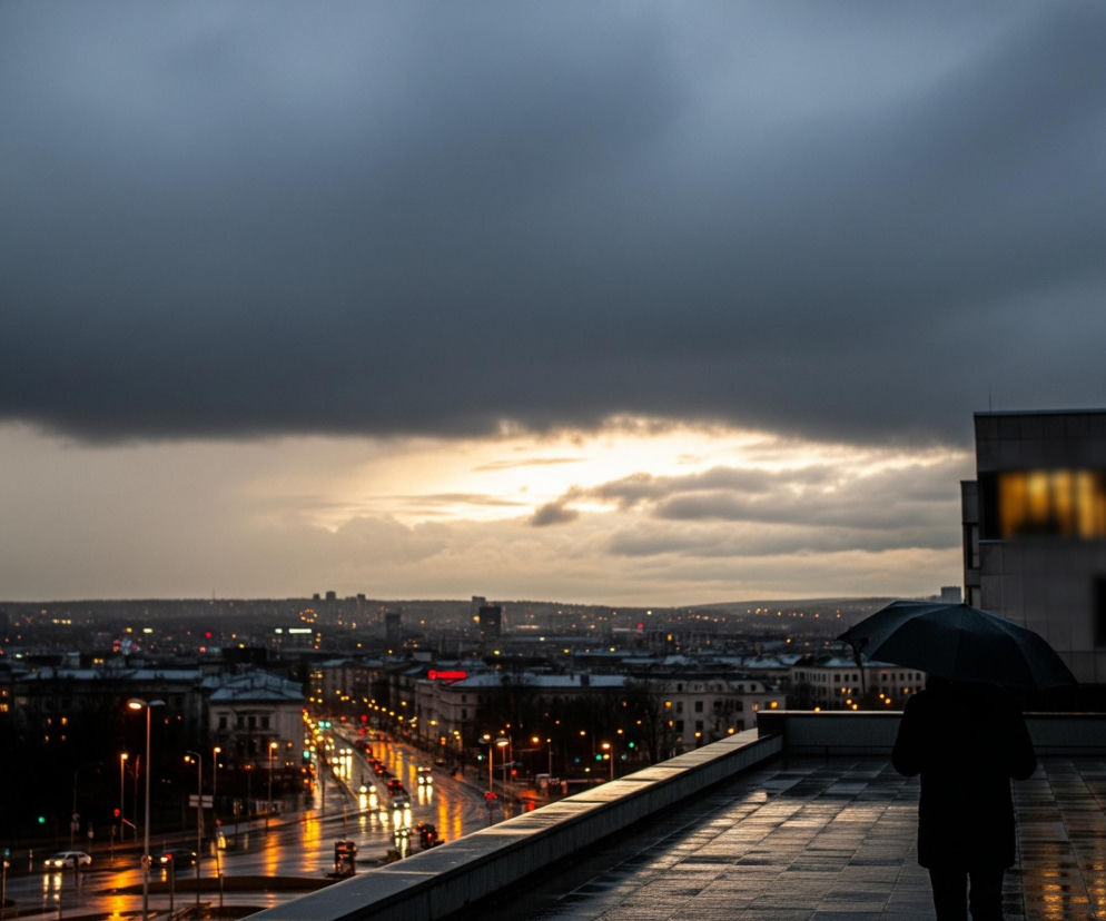 Osoba z parasolem, widoczna od tyłu w czarnej kurtce, stoi na mokrej, ciemnej posadzce tarasu lub dachu, skierowana w lewo, patrząc na panoramę miasta. Na horyzoncie rozciąga się miejski krajobraz z licznymi światłami budynków i ulic, których odbicia widać na mokrym asfalcie. Niebo jest zachmurzone, z wyraźnym pasem jaśniejszego, pomarańczowo-żółtego światła przy horyzoncie, wskazującym na zachód słońca lub świt, a nad nim dominują ciemne, szare chmury.
