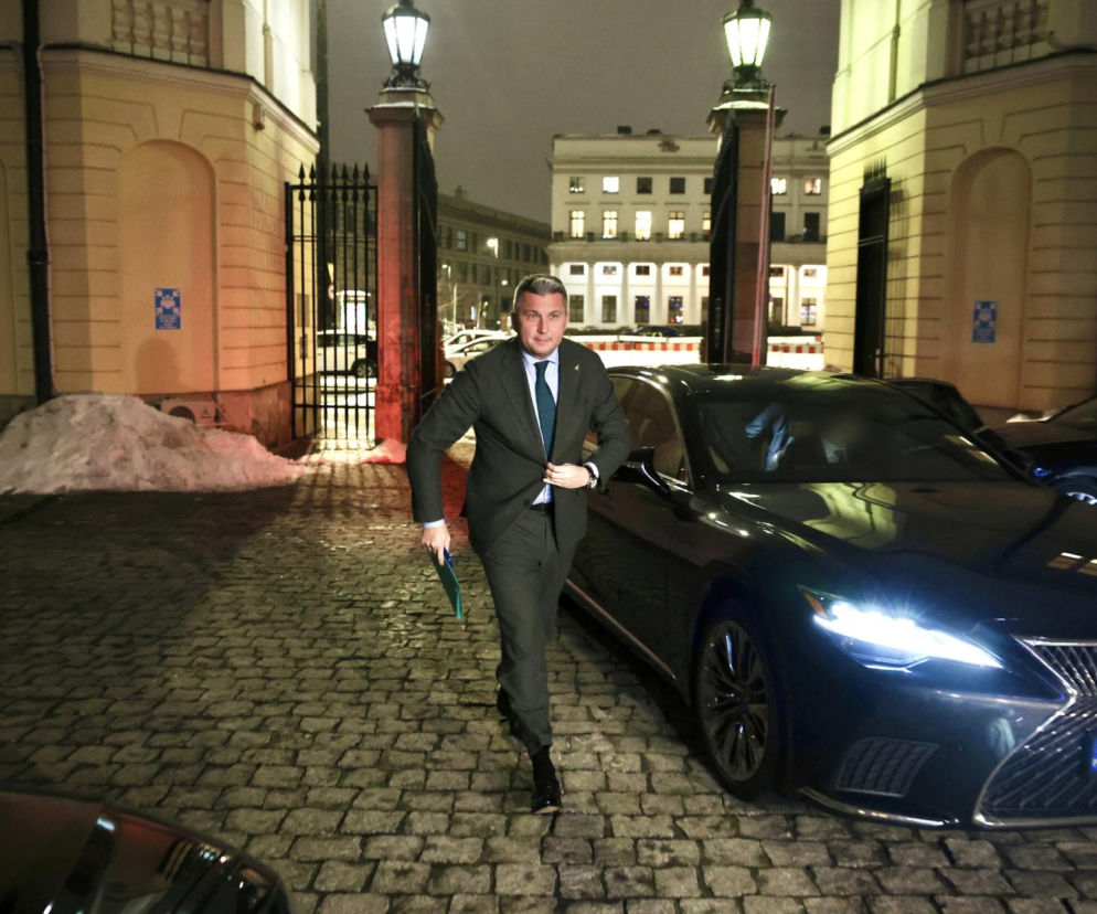 Radosław Piesiewicz, prezes PKOl, wychodzi z budynku w ciemnym garniturze z zieloną teczką, na tle zabytkowej bramy z latarniami i czarnego samochodu Lexus. Na Super Biznes możesz przeczytać o problemach PKOl ze sponsorami.