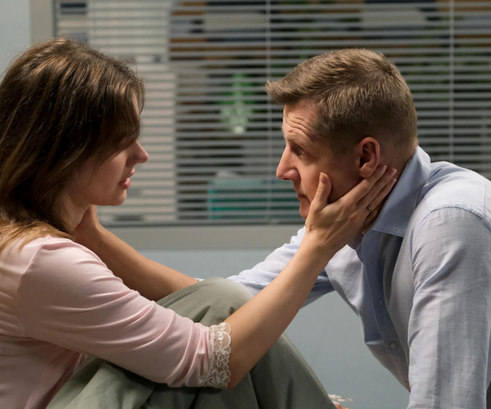 Dominika Kachlik (Franka) z długimi brązowymi włosami i w różowej bluzce, obejmująca dłońmi twarz Rafała Mroczka (Paweł) w niebieskiej koszuli, wpatrują się w siebie z troską, w tle rozmyte żaluzje. Zdjęcie ilustruje dramatyczne wydarzenia z serialu M jak miłość, o których przeczytasz na stronie Super Seriale.